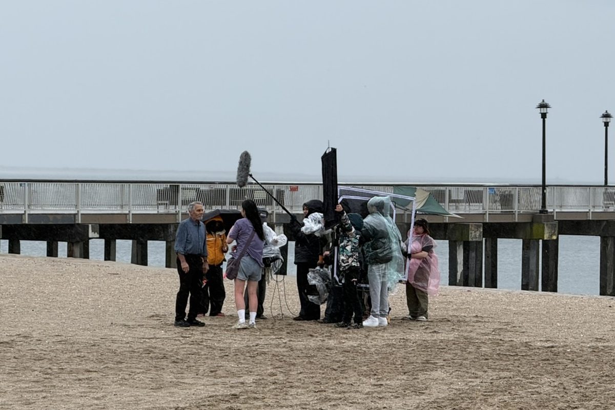 Production crew on a Coney Island beach in grey, rainy weather, wearing rain ponchos with a boom mic raised over the actors, pier visible in the background